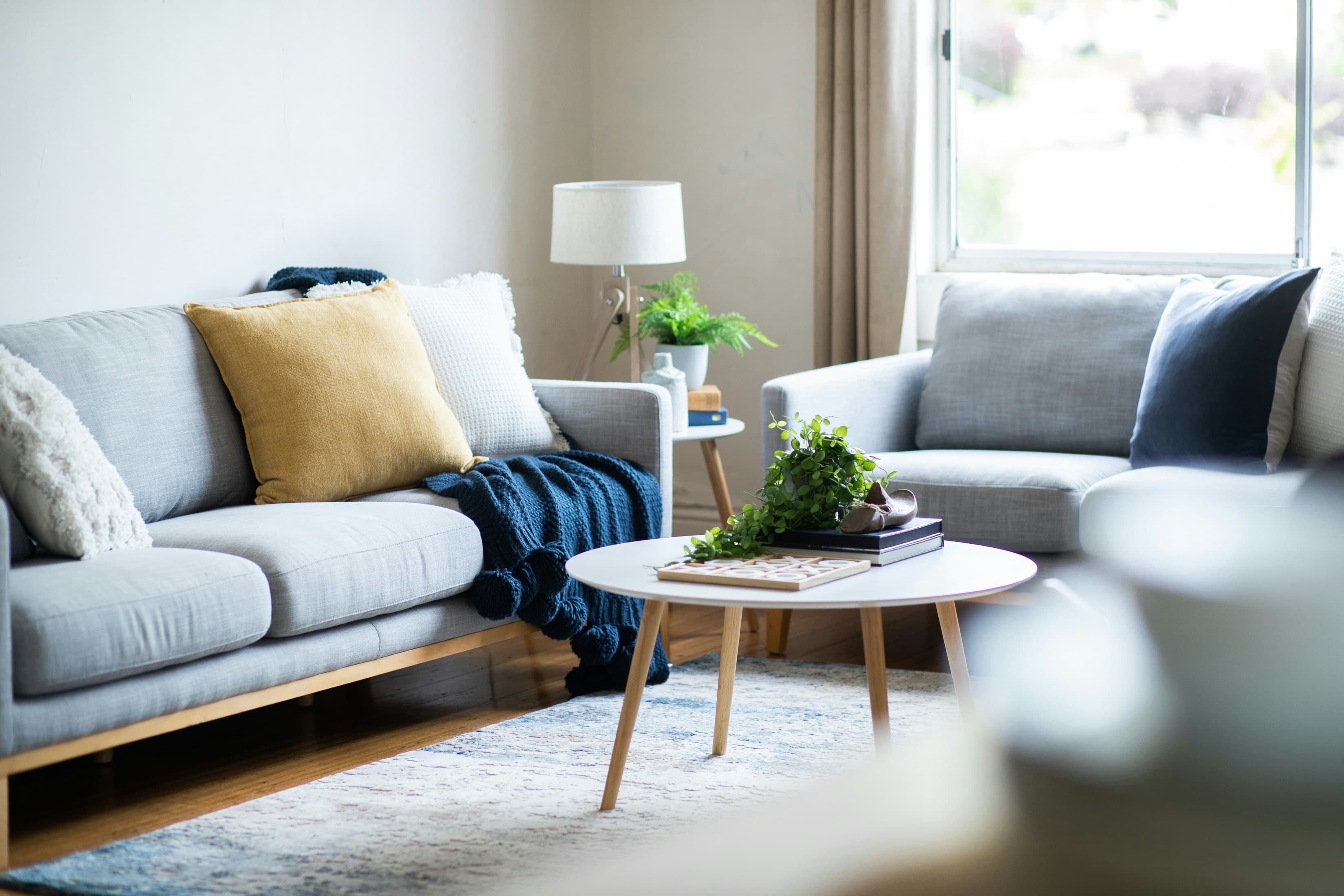 A warm, lived-in living room: two pale grey sofas, a mustard cushion and a navy knitted throw, a round timber coffee table and soft daylight from the window. An illustrative interior, not a Gather & Stay property.