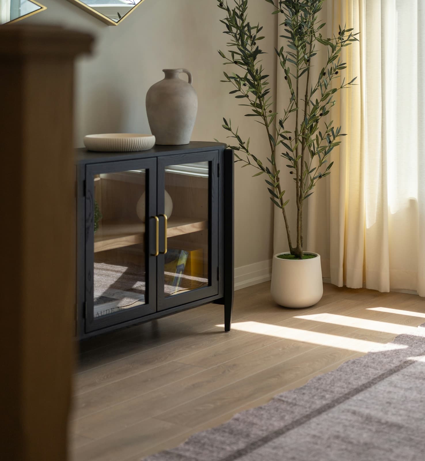 A quiet corner of a living room in afternoon sun: a dark glass-fronted cabinet with a stoneware jug and a shallow bowl, a potted olive tree against cream curtains, and window light falling in stripes across the timber floor. An illustrative interior, not a Gather & Stay property.