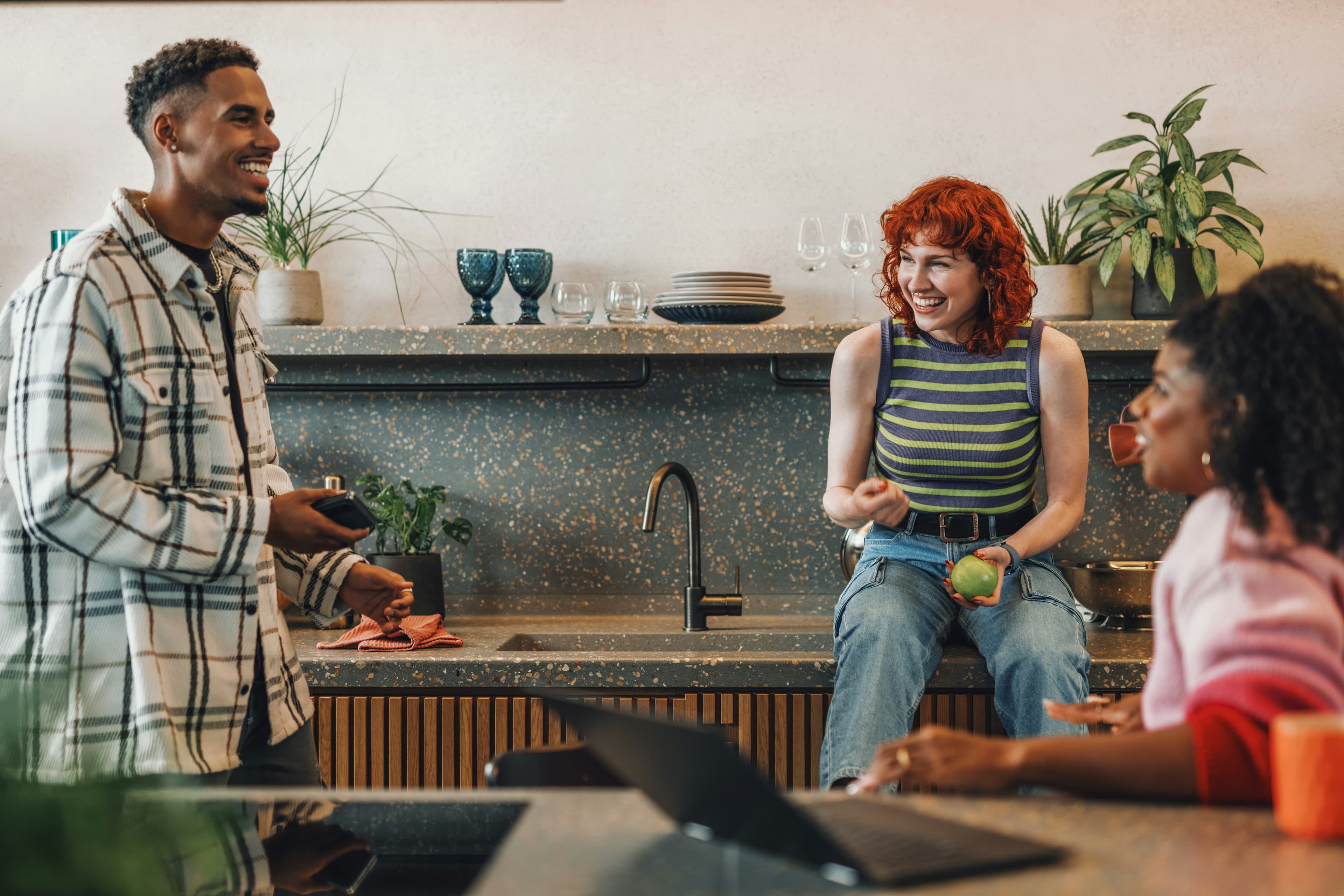 Three friends talking and laughing together in a kitchen, one perched on the terrazzo benchtop beside the sink, with shelves of glasses, plates and pot plants behind them