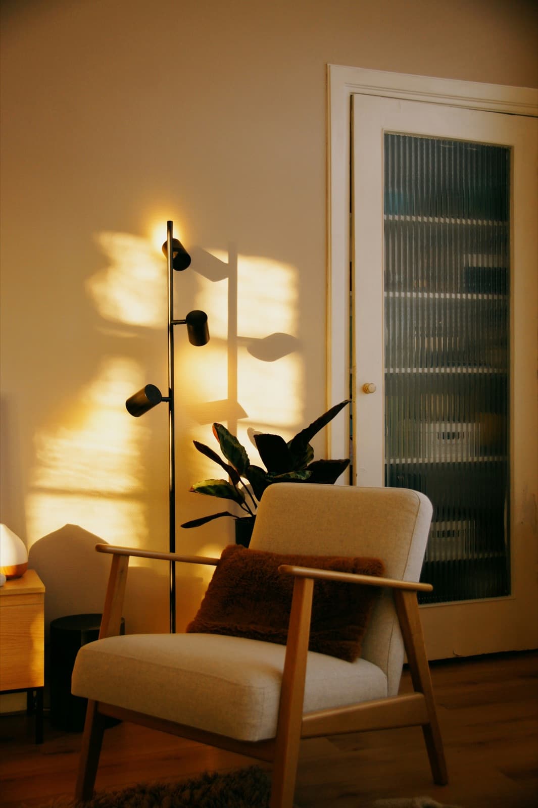 A cream armchair with timber arms and a rust sherpa cushion in the corner of a room, a floor lamp throwing warm light across the wall behind it