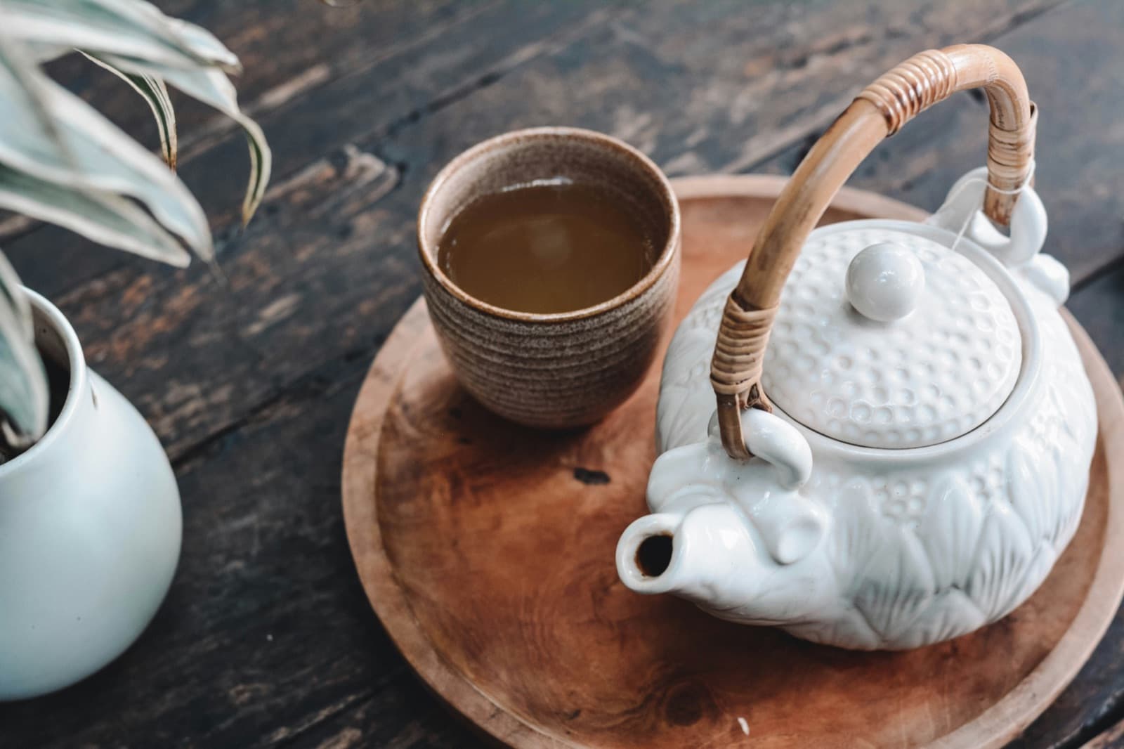 A white teapot with a cane handle and a glazed cup of tea on a round timber tray, on a weathered dark timber table