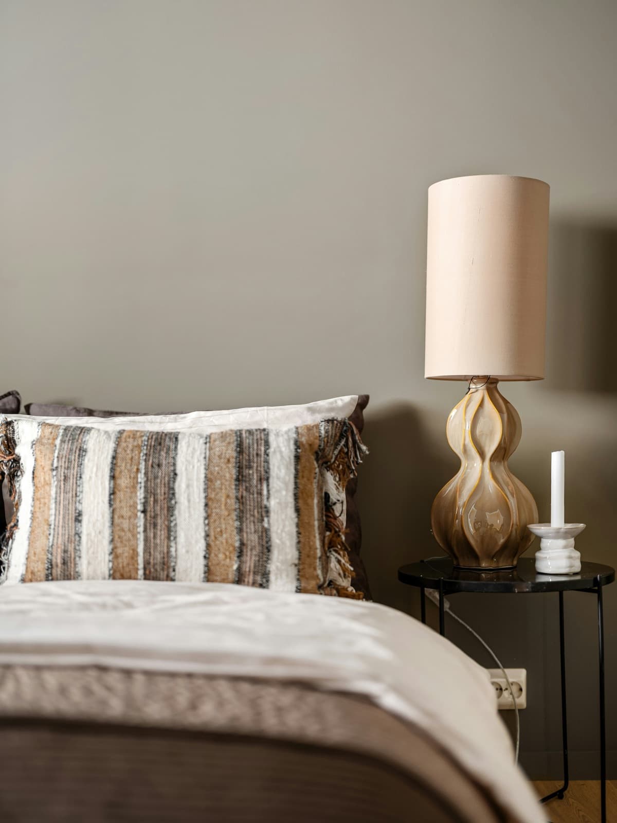 A made bed with white linen and a striped fringed cushion, beside a dark round table holding a sculptural glazed lamp and a single candle. An illustrative interior, not a Gather & Stay property.