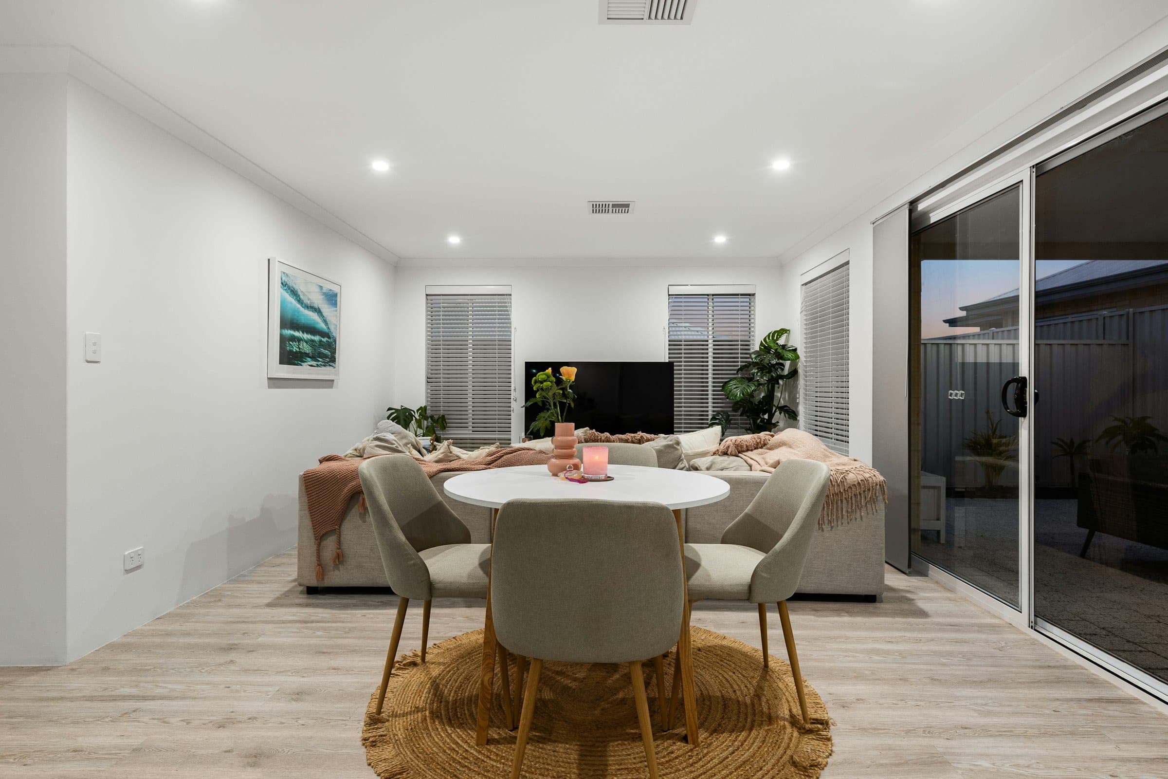 An open plan living and dining room in a single storey home, with a round table and armchairs on a level floor and a wide sliding door to a paved courtyard